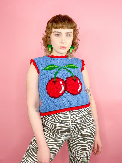 A woman posing wearing a bright blue crochet vest with a cherry on the front and red frilly edges, paired with zebra jeans and broccoli earrings.