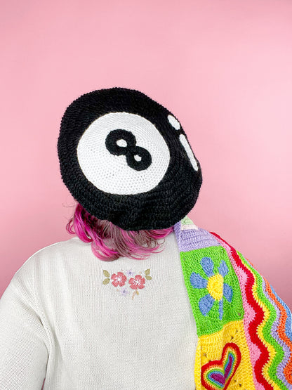 Back view of a woman wearing a eight ball beret in black with a granny style jumper and multicoloured scarf.