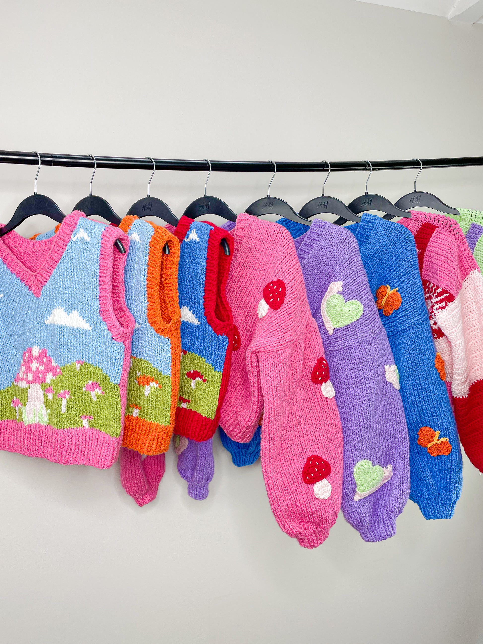 Three multicoloured hand knitted vests, three knitted cardigans and two crocheted jumpers hung up together on a metal clothes rack in front of a white background.