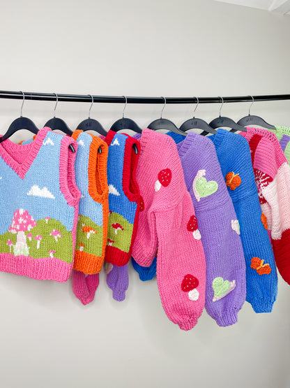 Three multicoloured hand knitted vests, three knitted cardigans and two crocheted jumpers hung up together on a metal clothes rack in front of a white background.