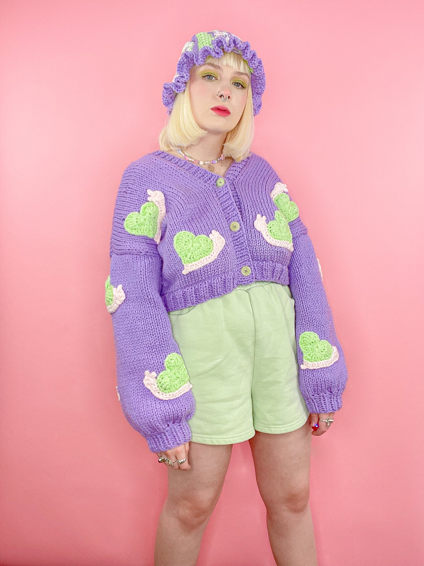 A woman posing wearing a lilac knitted cardigan with cream and mint green snails sewn all over it with a lilac and mint green crochet bucket hat and mint green shorts.