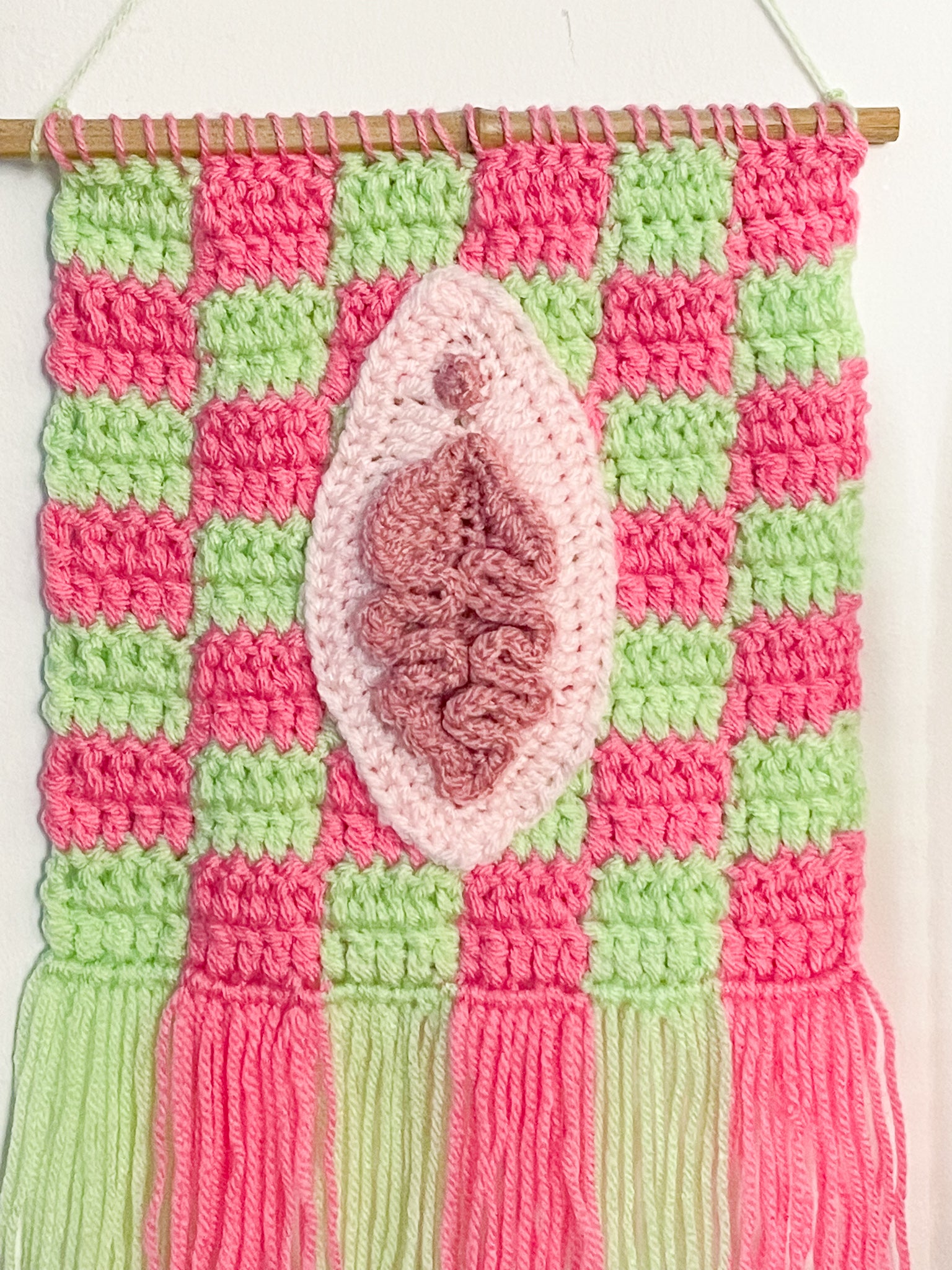 Close view of a crochet tapestry with a check background and appliqué that resembles a vulva in cream and pink, attached to a wooden dowel rod.