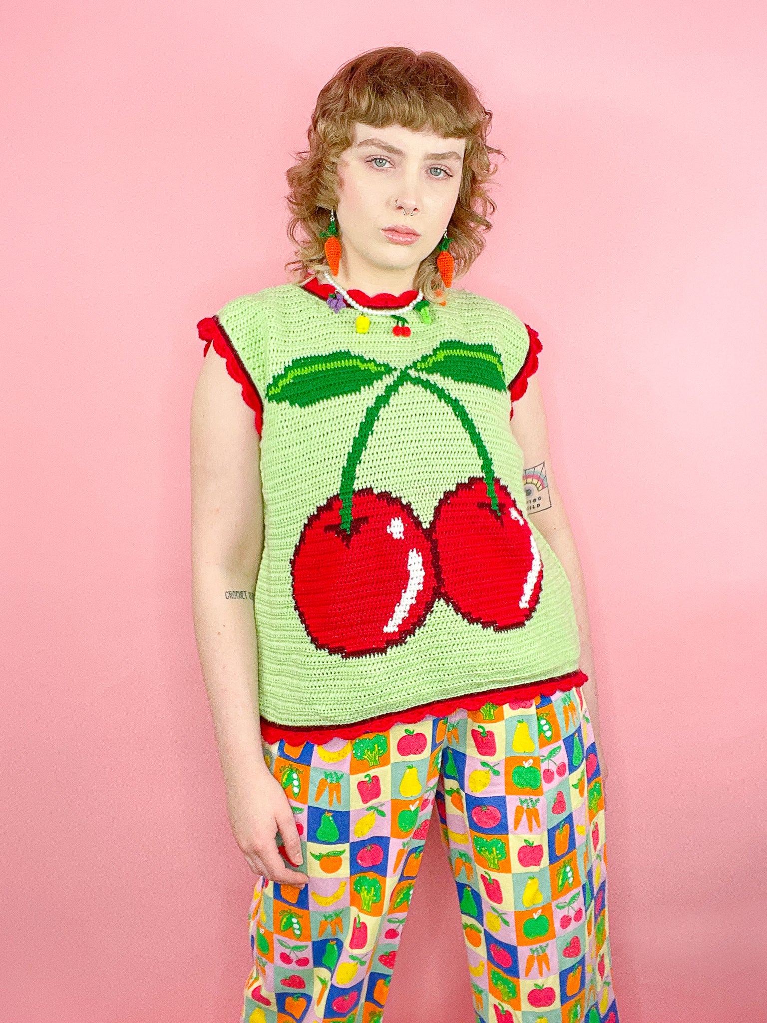 A woman wearing a green vest with a crochet cherry on the front paired with fruit and veg check pants, carrot earrings and a fruity choker.