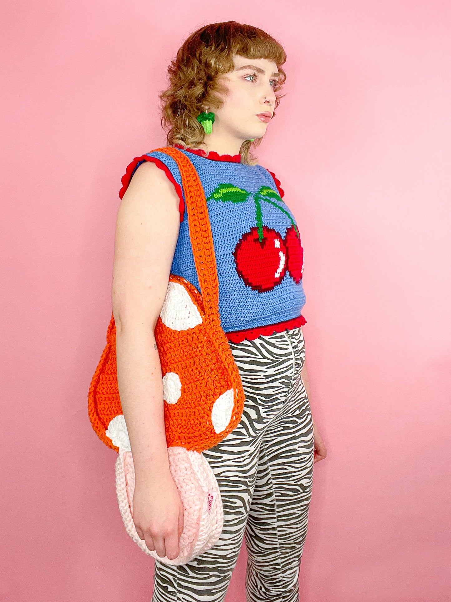 Side view of woman wearing a toadstool bag in orange with a blue vest with a red cherry on it and zebra print jeans.