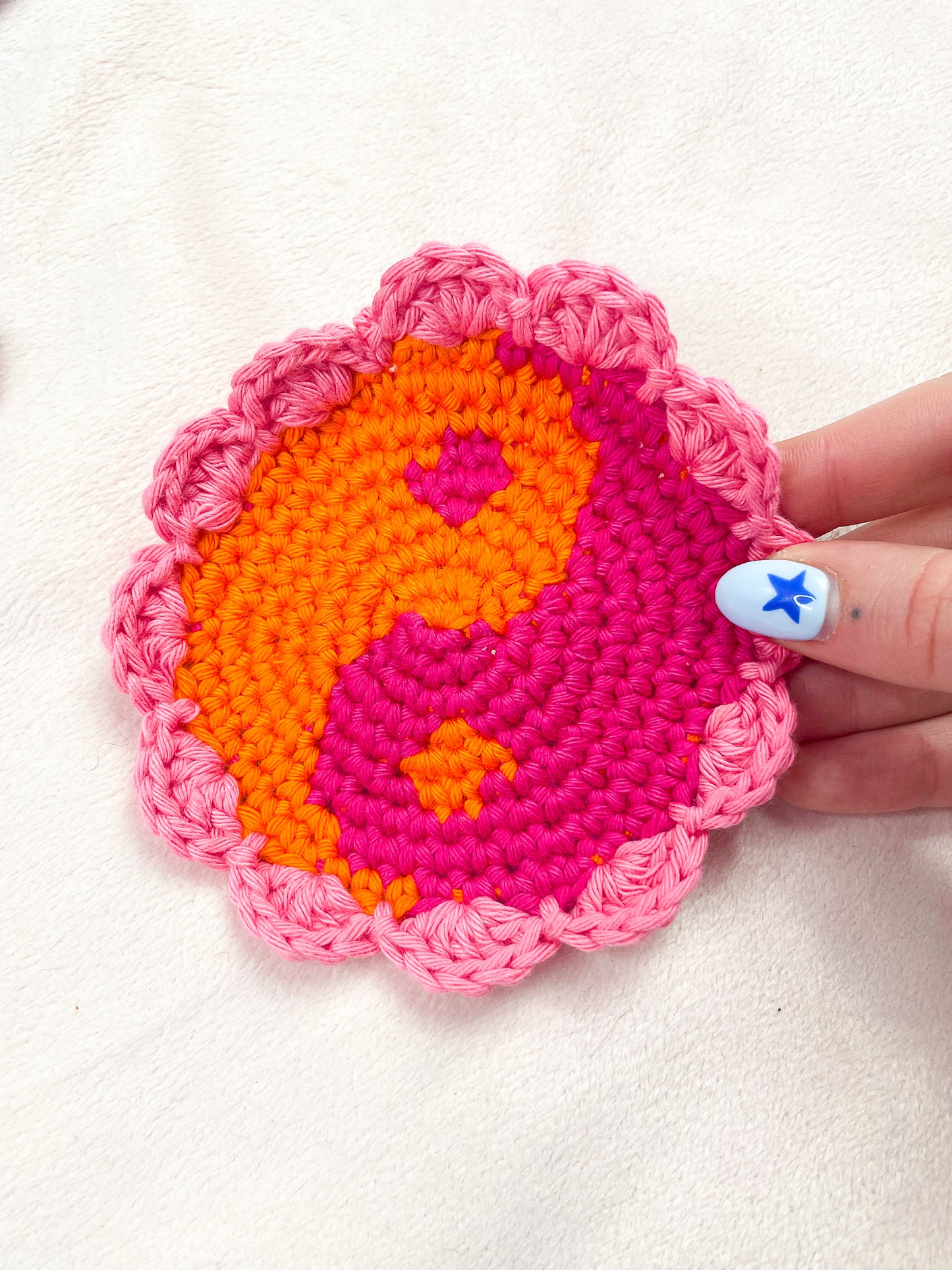 A hand holding a crochet coaster that looks like a yin yang in bright orange and pink with a light pink scalloped edge.