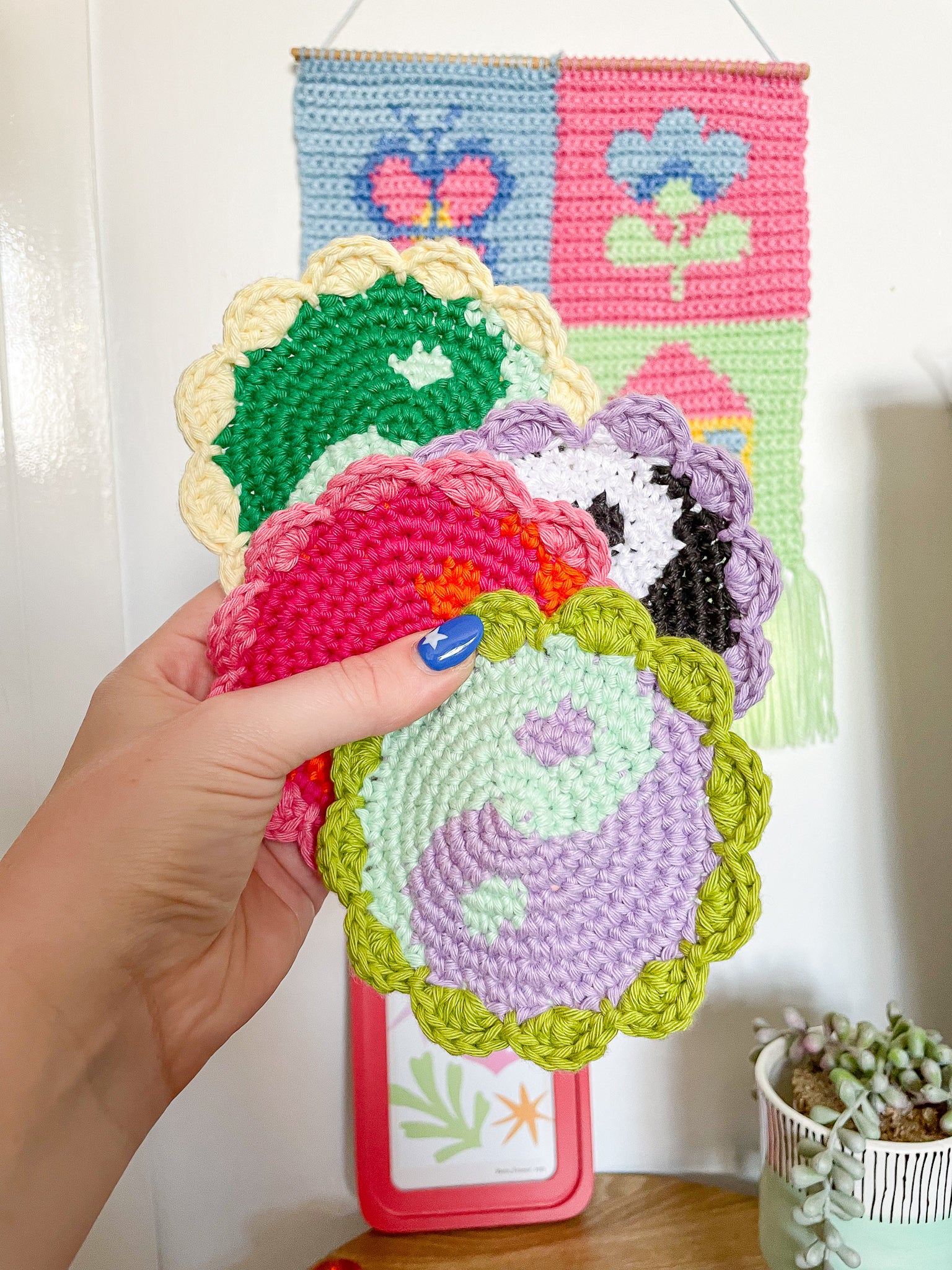 A hand holding four crochet coasters with yin and yang designs in different colours held in front of a white wall with decor around it.