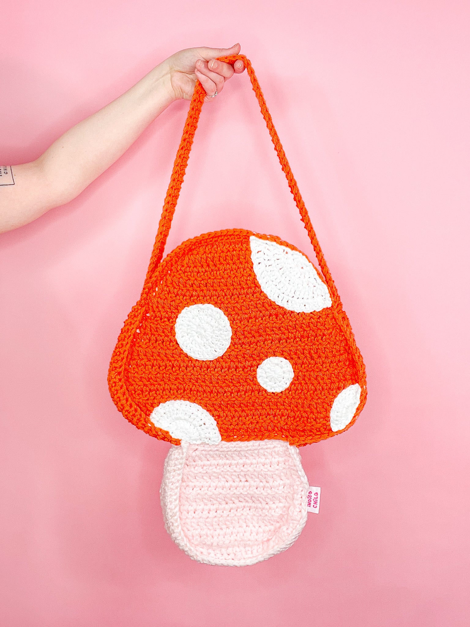 A hand holding an orange crochet bag shaped like a mushroom against a pink background.