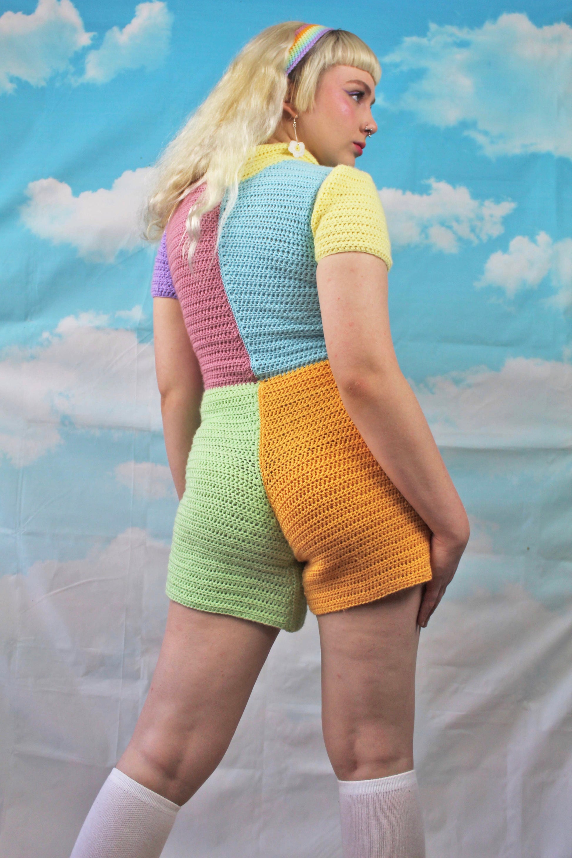 Back below view of woman posing wearing a crocheted playsuit in patchwork pastel colours with white knee high socks and a pastel rainbow headband.