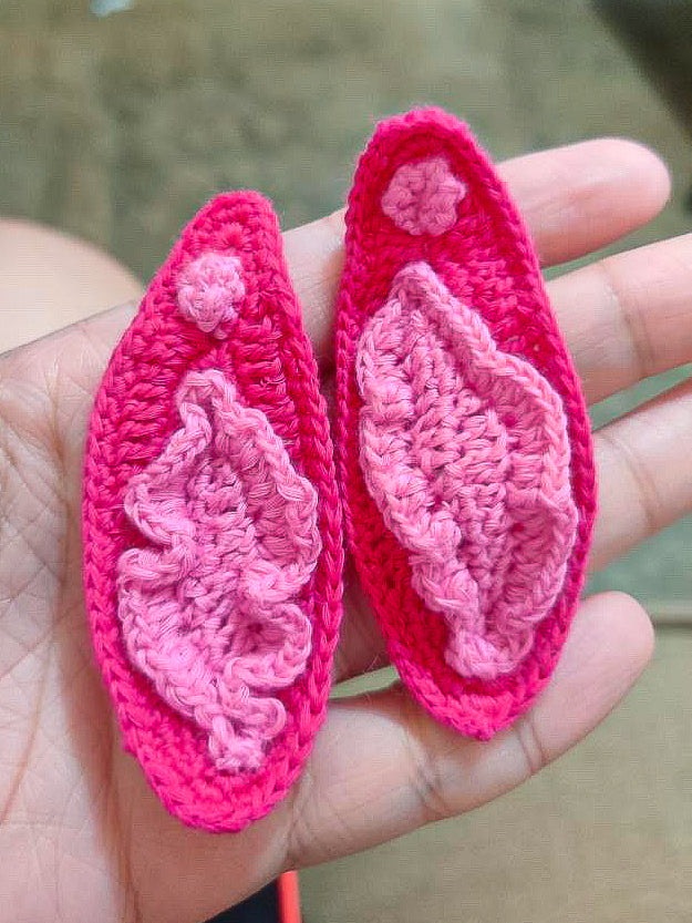 A hand holding a pair of  crocheted earrings inspired by vulvas with light pink detailing and a bright pink base.