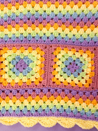 Close up of a pastel rainbow skirt with a striped design and crochet granny squares in the middle lying on a purple background.