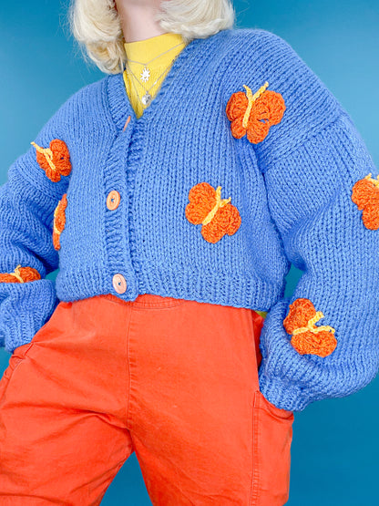 Close up of a woman wearing a knitted cardigan in royal blue with orange butterflies all over and orange buttons with a yellow turtleneck and orange overalls.