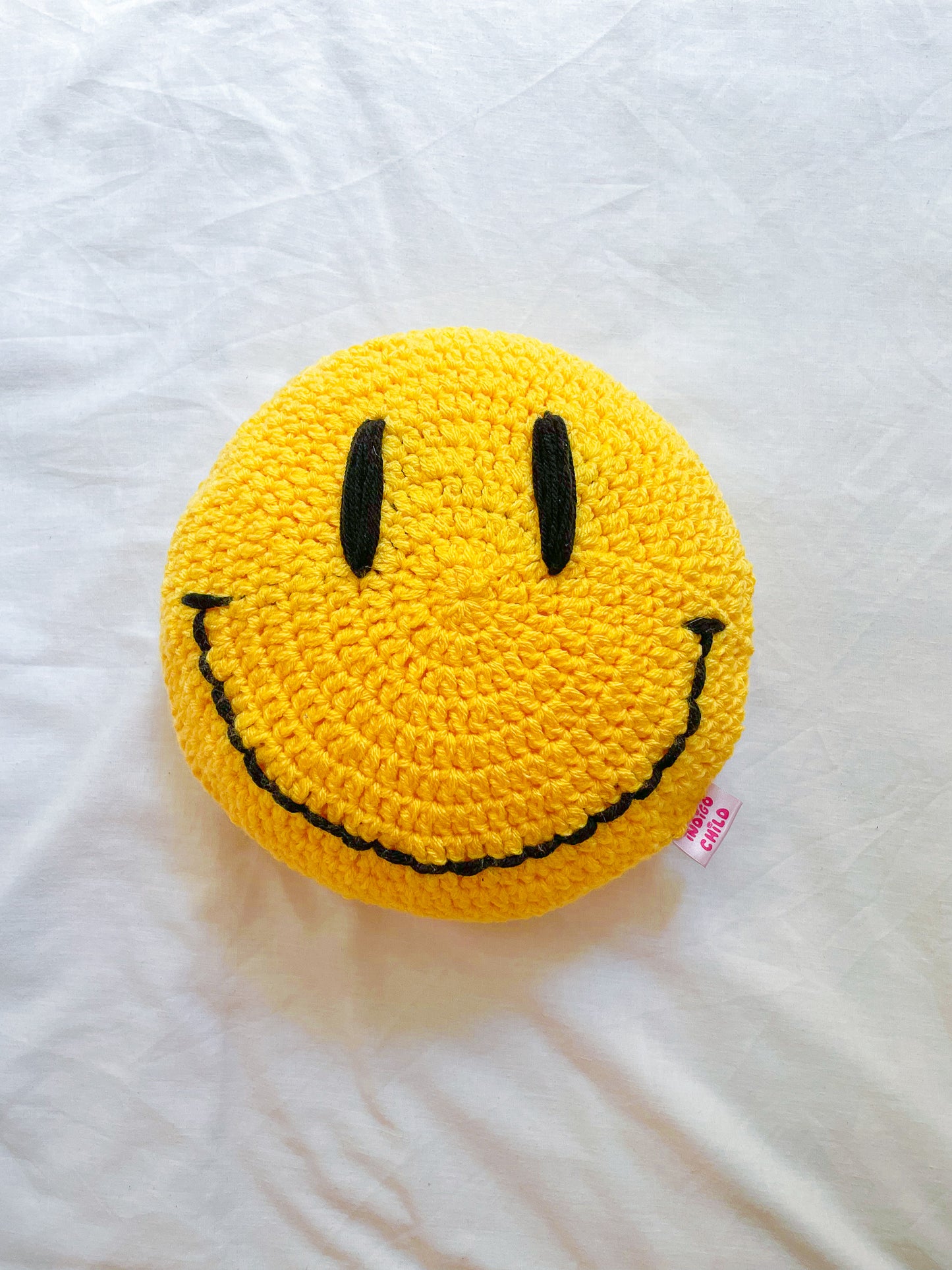 A yellow circle pillow with a happy face on it lying flat on a white background.