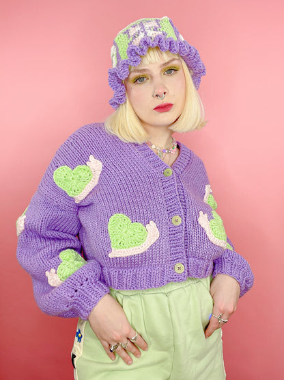 Close view of a woman posing wearing a lilac hand knit cardigan with cream and mint green snails on it with a crochet bucket hat and matching green shorts.