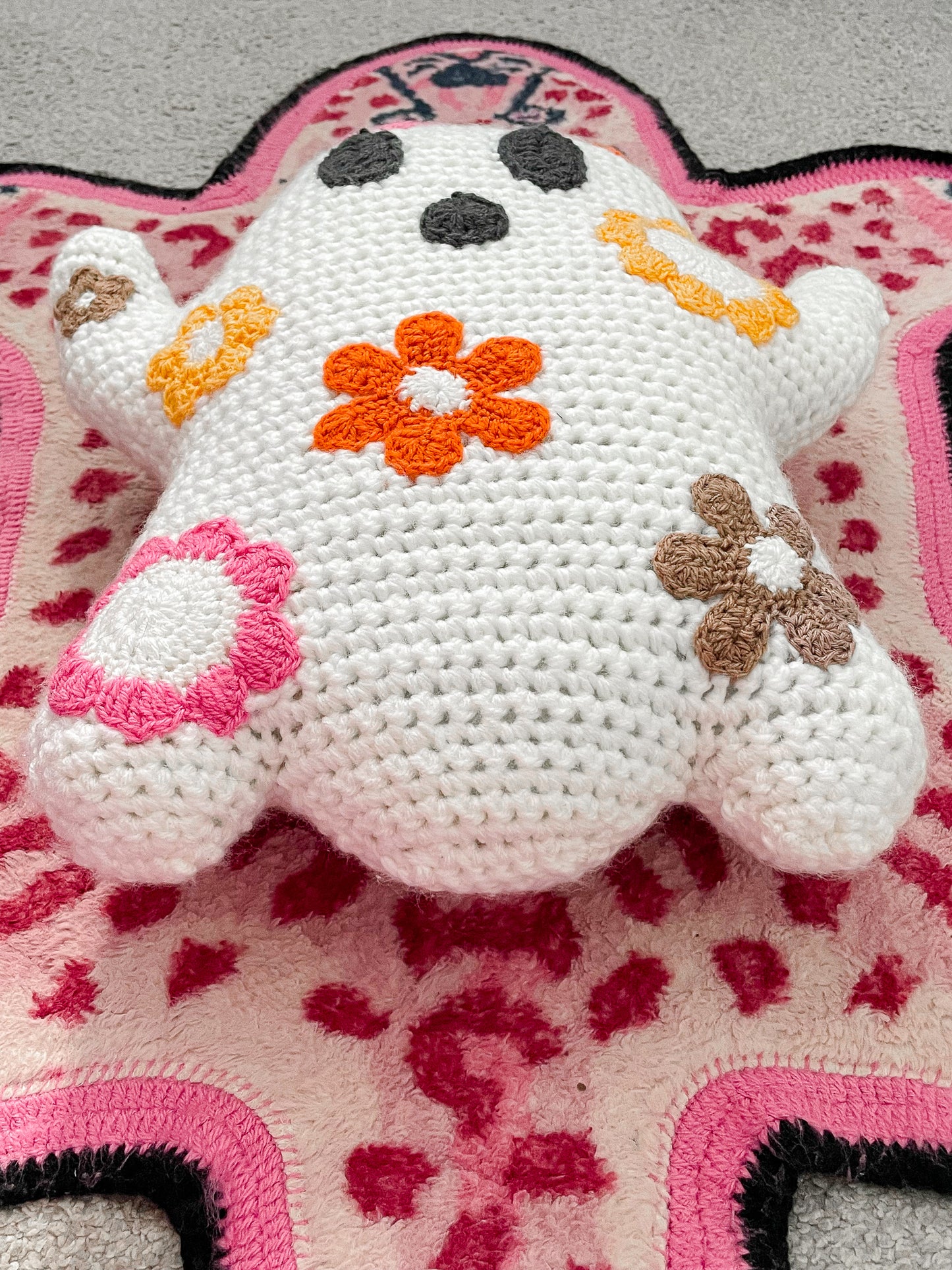 Below angle of a ghost shaped pillow with flowers attached lying on a pink leopard rug showing the crochet stitches.