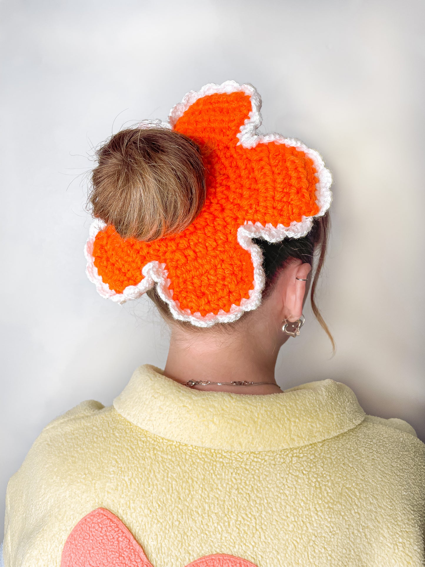 Side view of a woman wearing a crochet flower scrunchie in orange with a matching yellow fleece with a pale orange flower on the back.