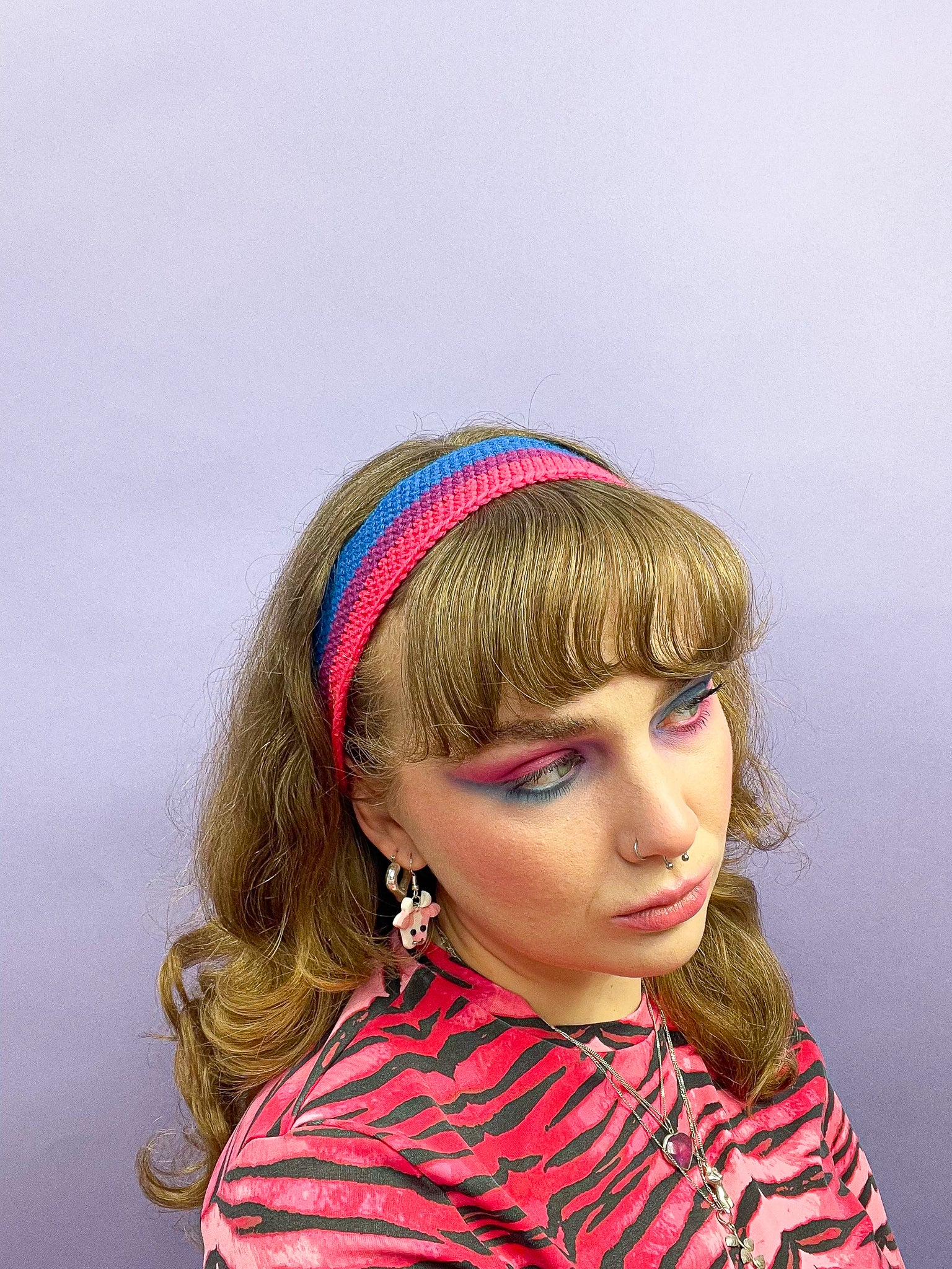 Side view of a woman posing wearing a bisexual flag headband (blue, purple, pink), with a matching pink zebra top and blue and pink eyeshadow.