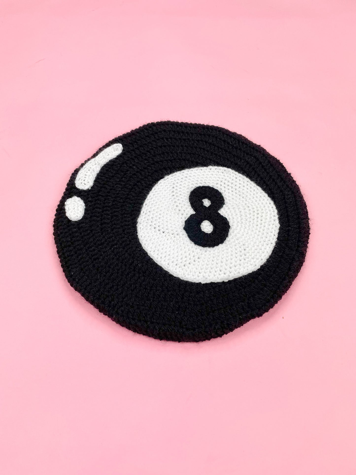 Flat lay angle view of a black crochet beret resembling an eight ball lying on a pink background.