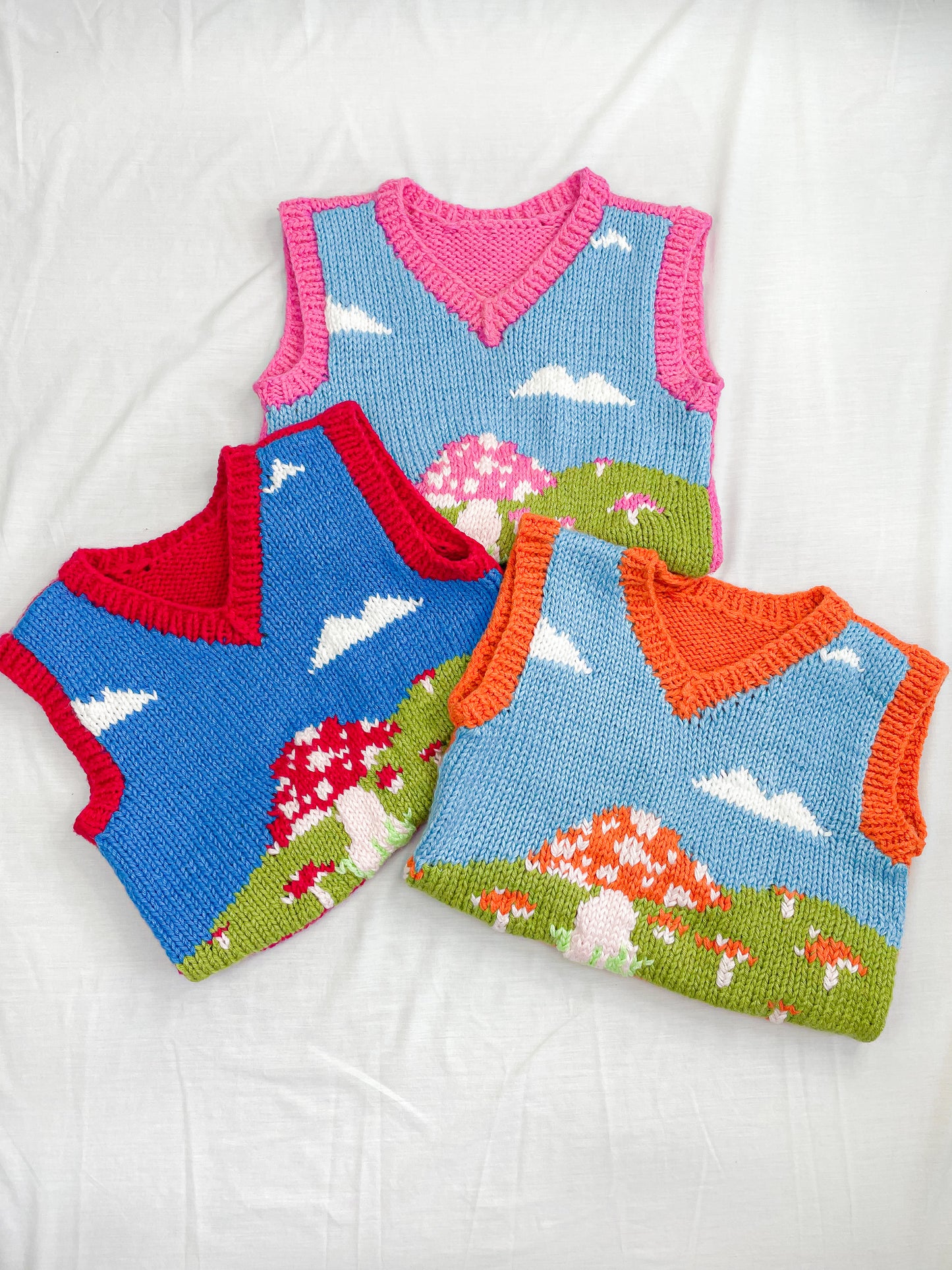 Flat lay of three knitted vest with mushroom field designs in pink, red and orange, folded in half and lying on top of each other on a white background.