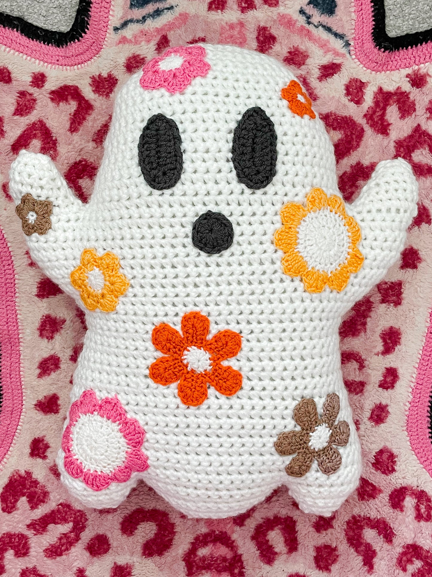 Flat lay of a crochet ghost pillow with flowers all over it lying on a pink leopard print rug.