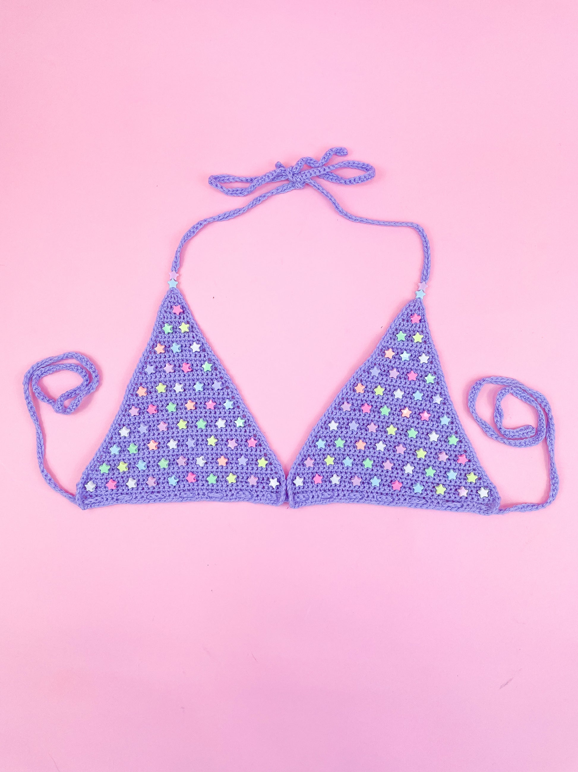 Flat lay of a lilac triangle bralette with star beads on the cups lying on a pink background.