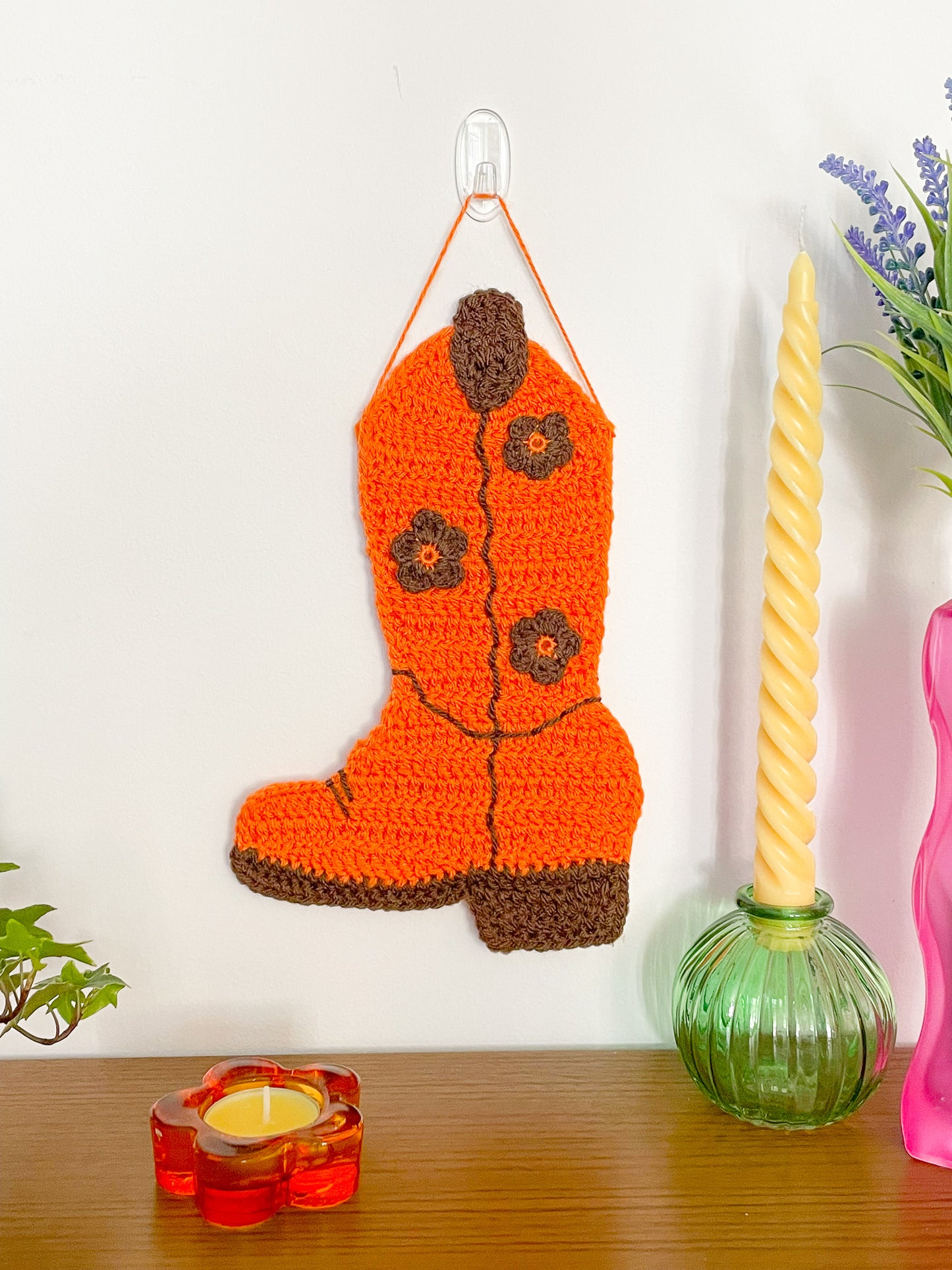 An orange cowboy boot wall hanging with brown stitches and details hanging on a white wall with a wooden table beneath it and candles and plants around it.