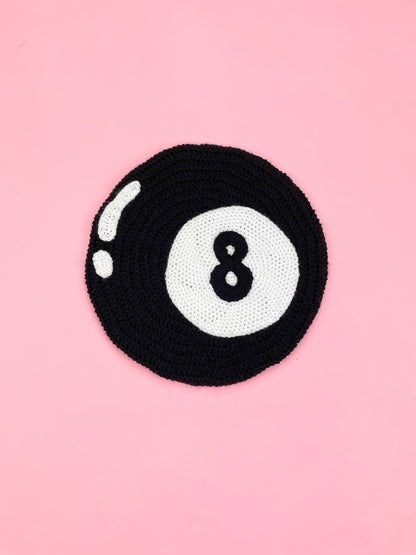 Flat lay of a black beret with an eight ball design lying on a pink background.