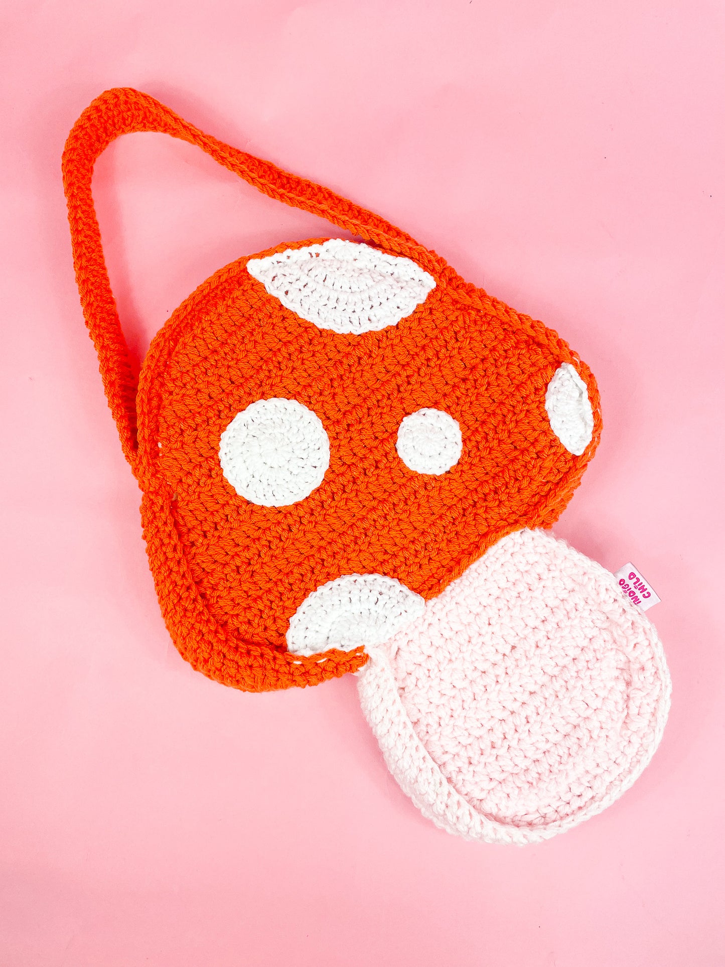 A mushroom shaped crochet bag in orange lying flat on a pink background.