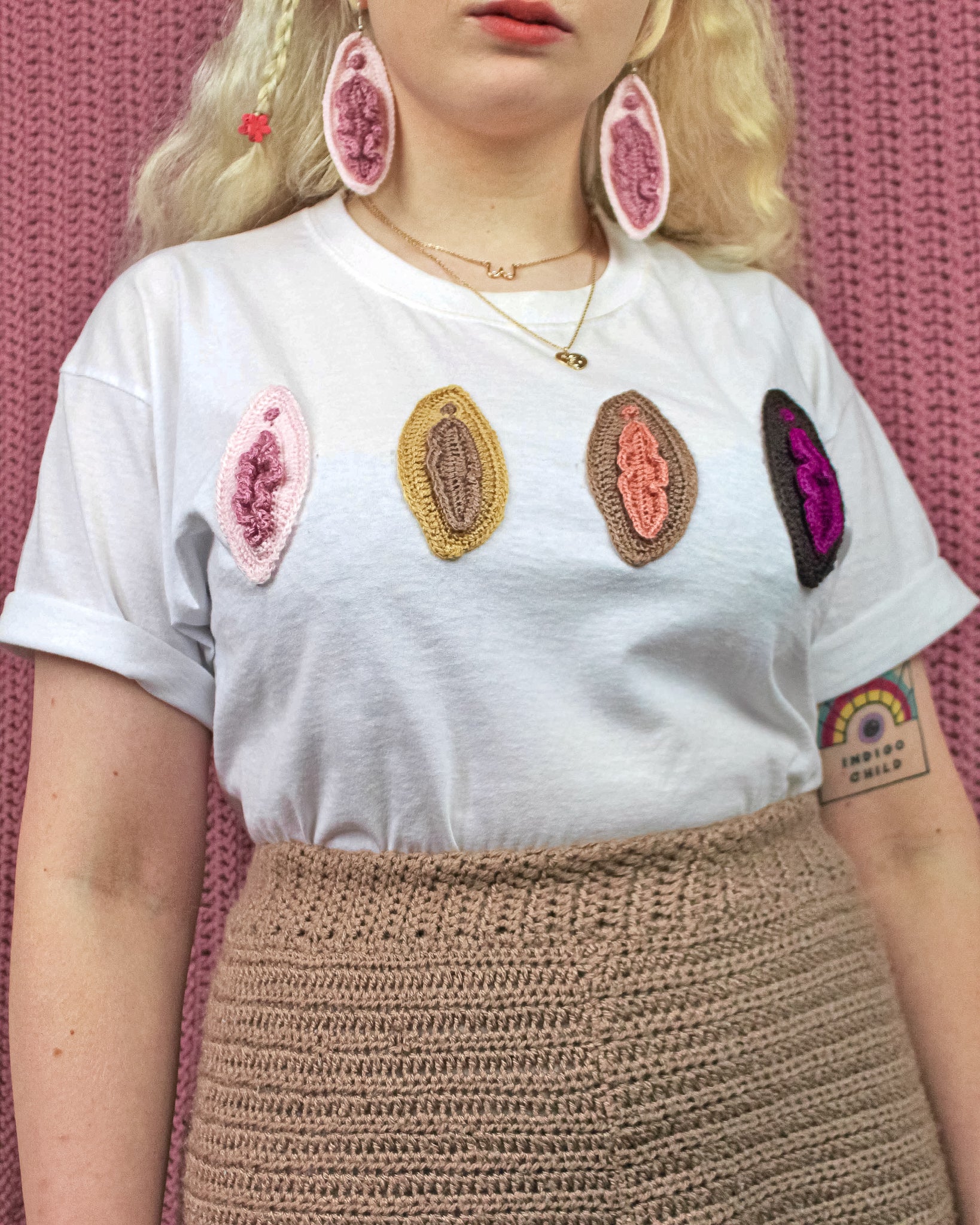 Below close view of a woman wearing a white shirt with crochet appliqué vulvas on the front with brown trousers and matching vulva earrings.