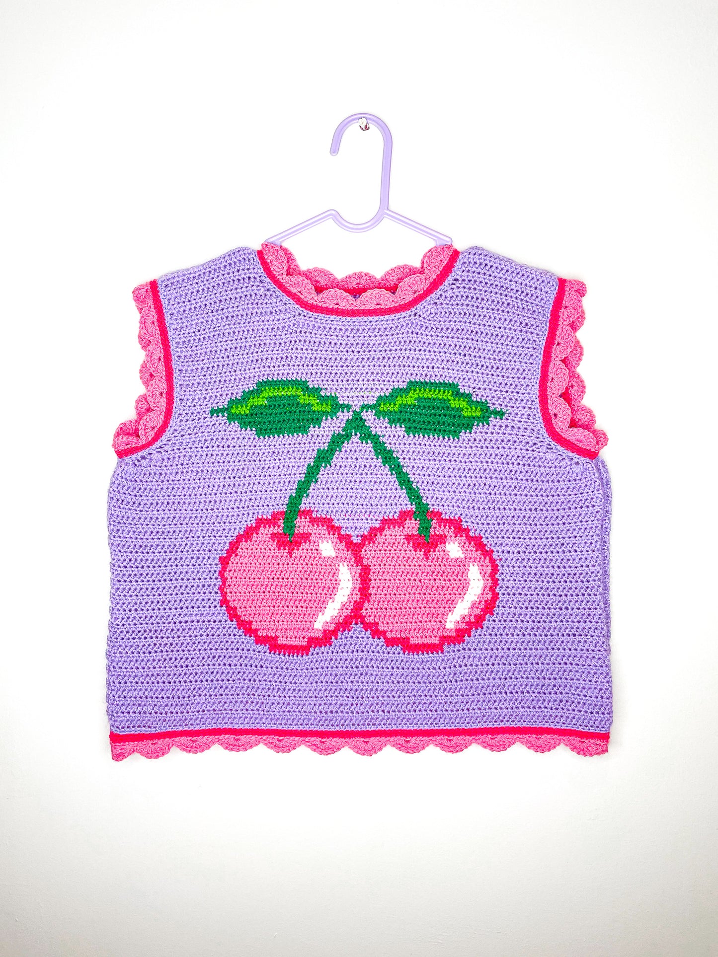 A lilac crochet vest with a pink cherry on the front and scalloped hemlines hung on a white wall.