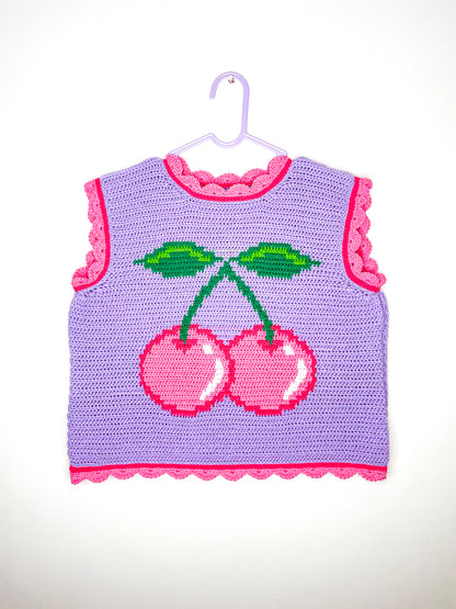 A lilac crochet vest with a pink cherry on the front and scalloped hemlines hung on a white wall.