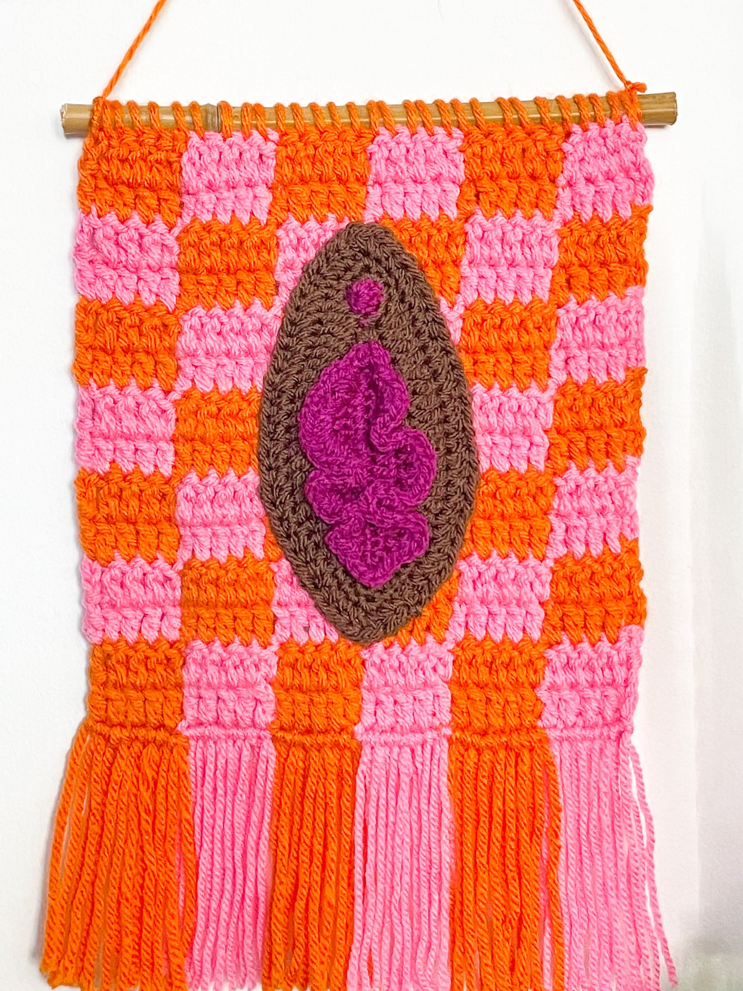 Close view of an orange and pink tapestry with a dark brown vulva attached to the front with orange and pink tassels attached to the edge.