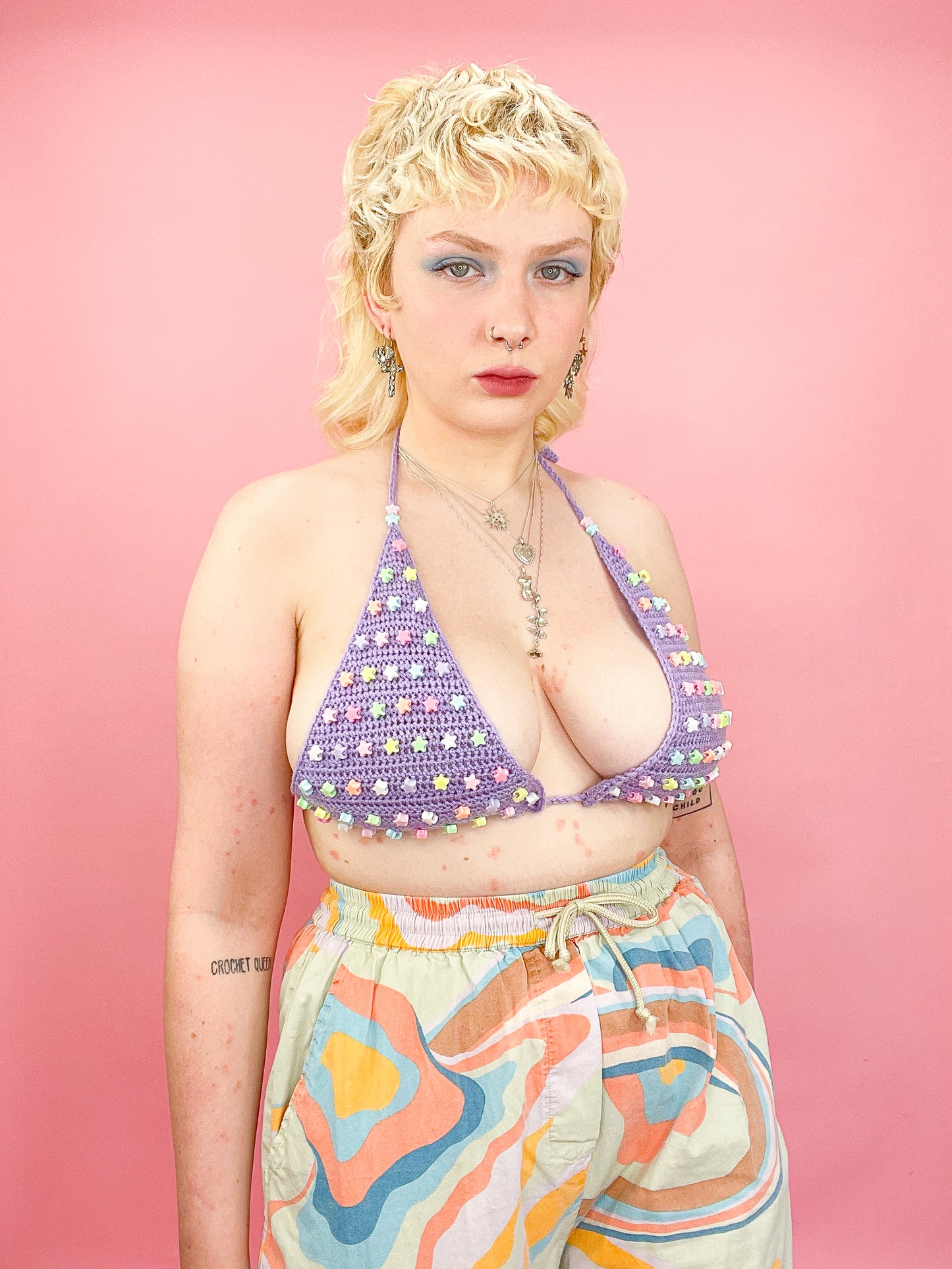 A woman posing wearing a lilac crochet bralette with pastel star beads on the cups with patterned trousers and silver jewellery.