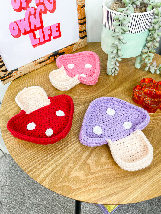 Three mushroom ring trays in pink, red and purple lying together on a wooden coffee table with a picture frame, plant and candle holder.