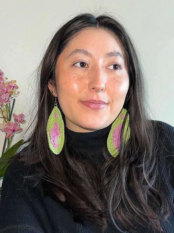 A woman wearing a pair of vulva earrings in brown and pink standing next to a pink orchid.