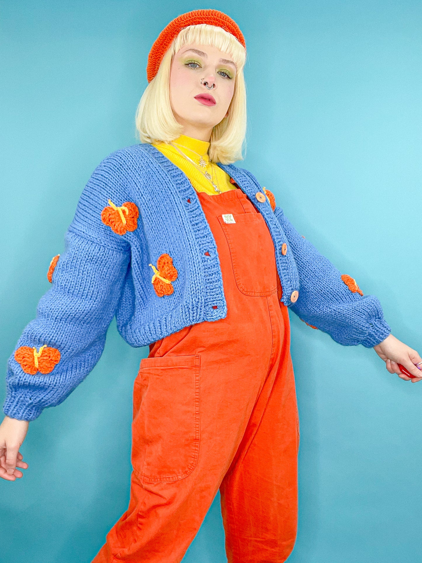 A woman posing wearing a blue hand knitted cardigan with orange crochet butterflies all over it paired with an orange beret, yellow turtleneck and orange overalls.