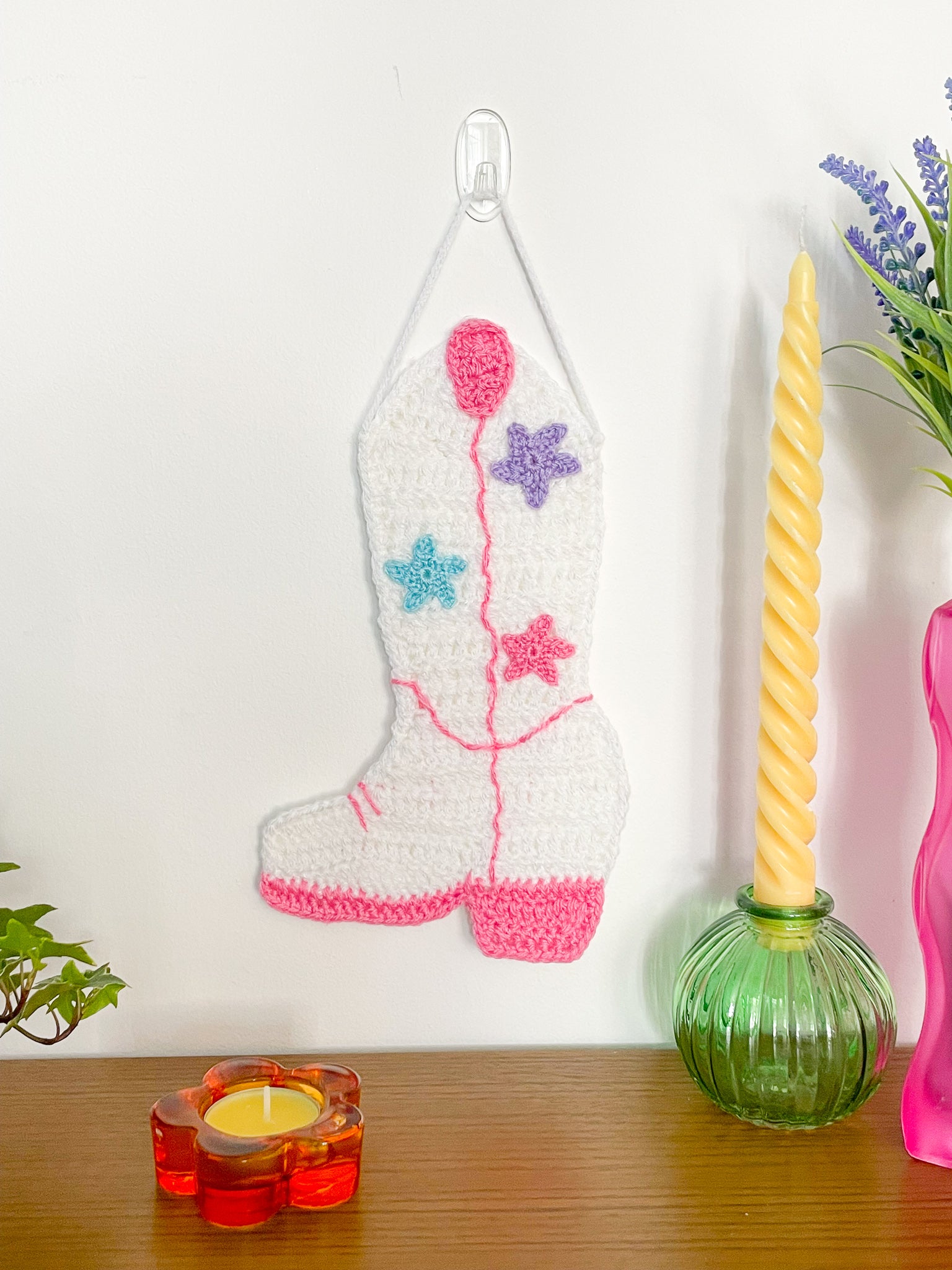 A white cowboy boot wall hanging with pink stitches, a pull strap and hell and blue, pink and lilac crochet appliqué stars hanging on a white wall with home decor around.