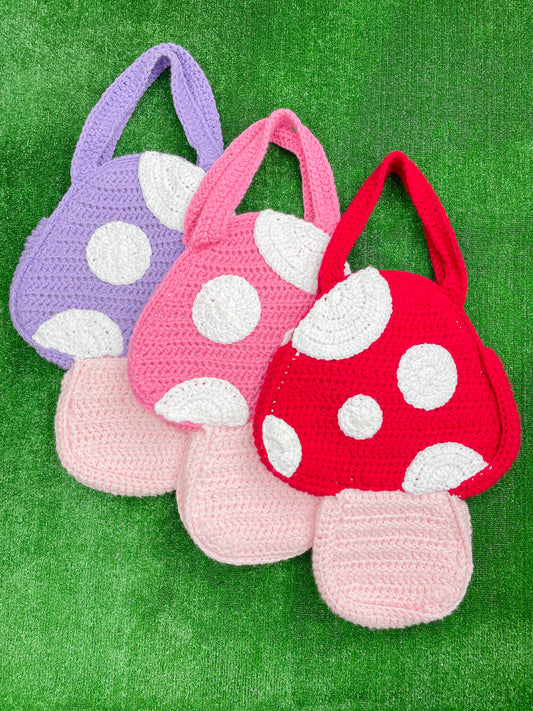 Three mushroom shaped tote bags in lilac, pink and red laying on top of each other in a row on grass.