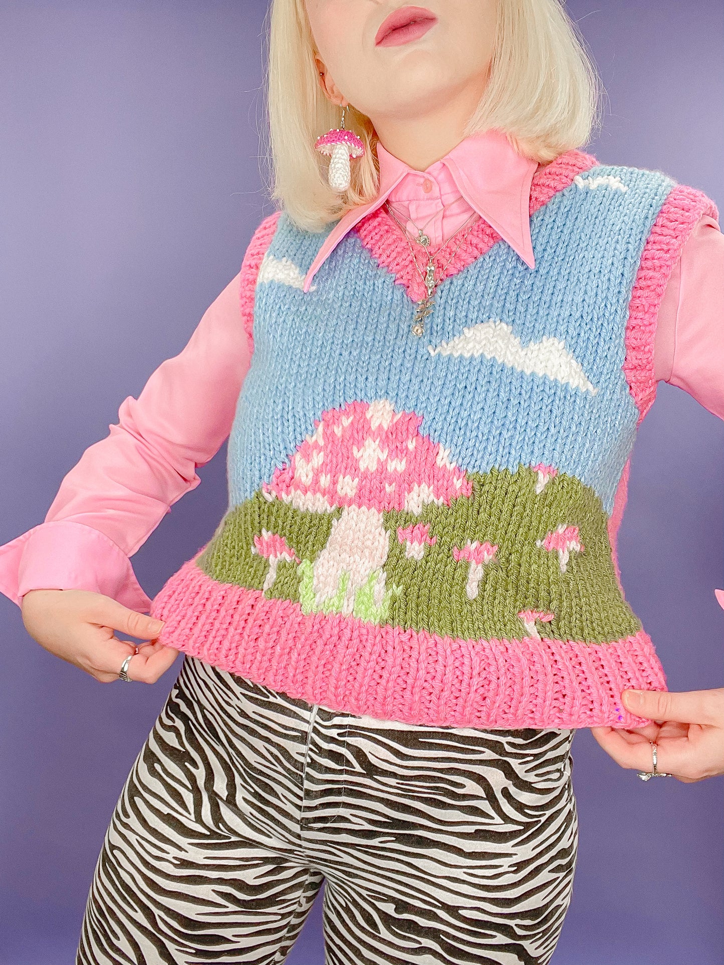 Close up of a woman wearing a hand knit vest top with a mushroom fields motif and pink edges, paired with a pink 70's shirt and zebra print jeans.