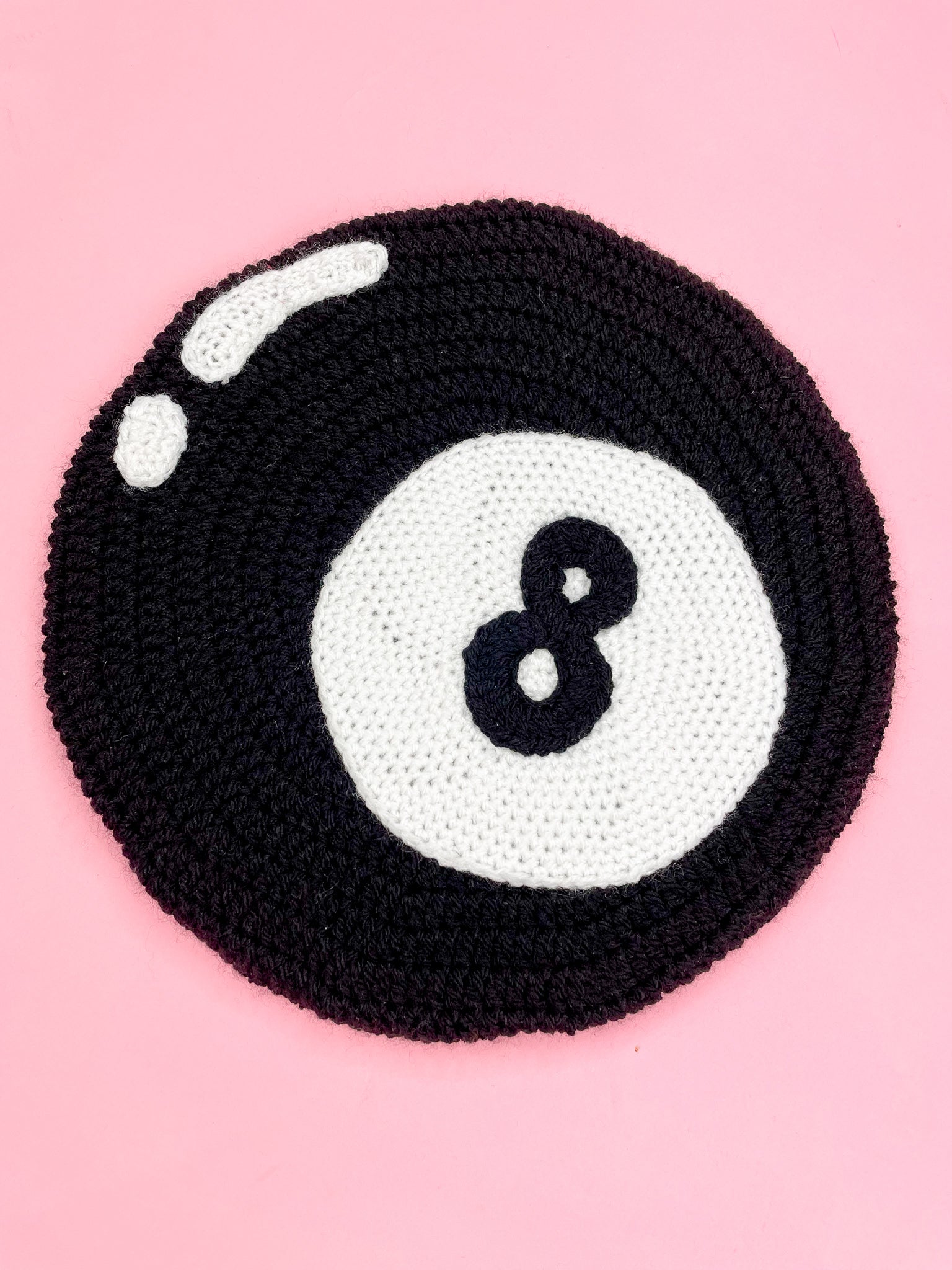 Flat lay close up of a black eight ball beret with crochet appliqué details to make the design lying on a pink background.