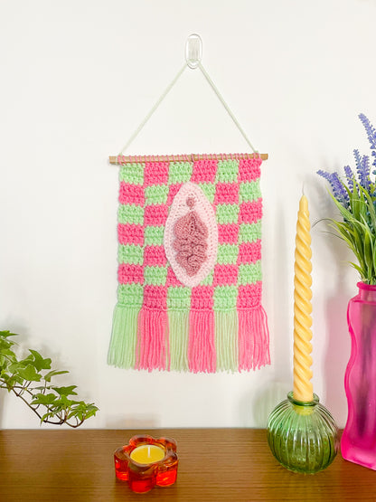 A crochet wall hanging with a check pink and mint green background and appliqué art resembling a vulva on the front with some candles and plants around it.