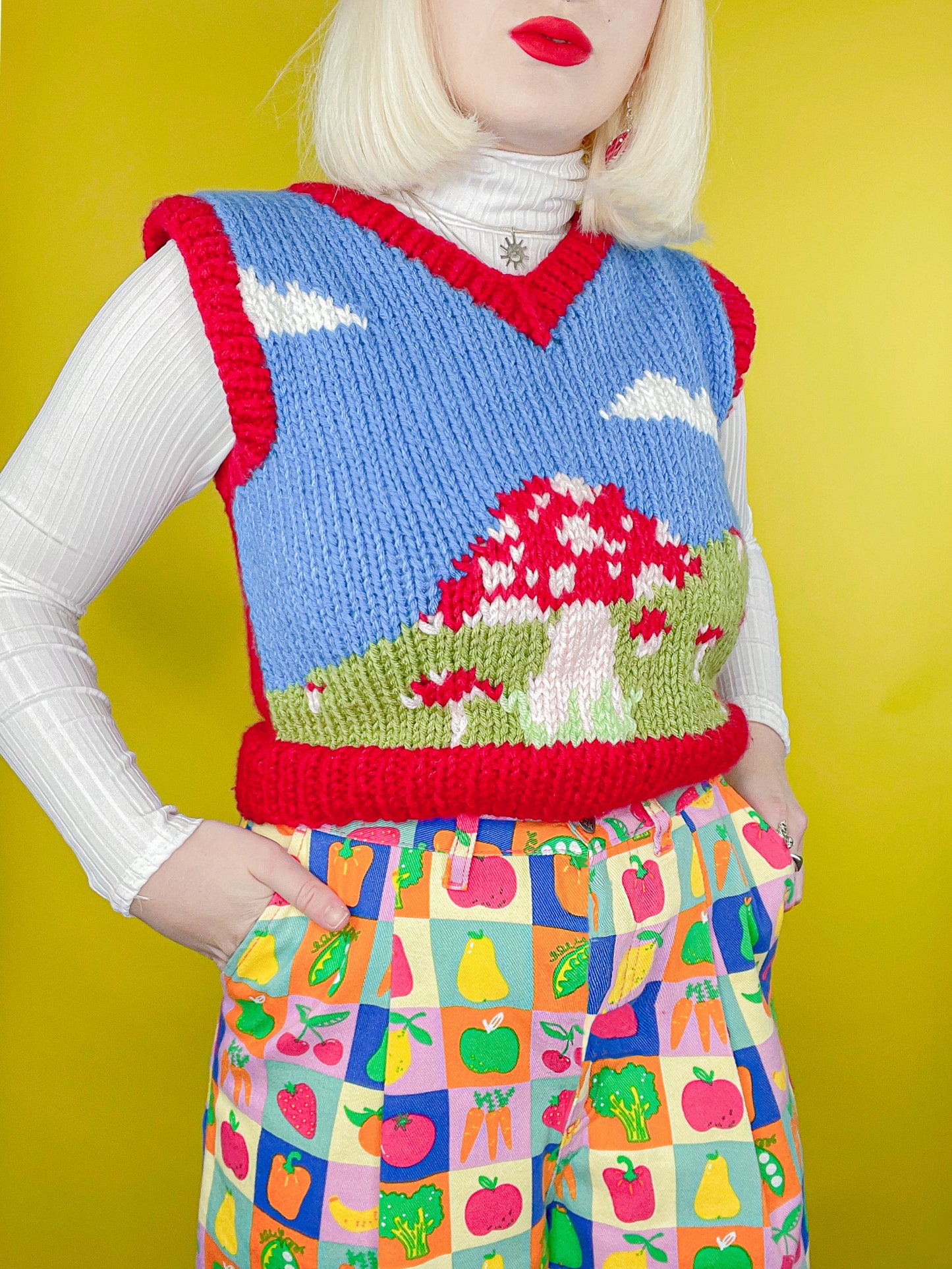 Close up of woman wearing a knitted vest top with a mushroom fields and clouds motif in dark blue and red with patterned jeans standing in front of a yellow background.