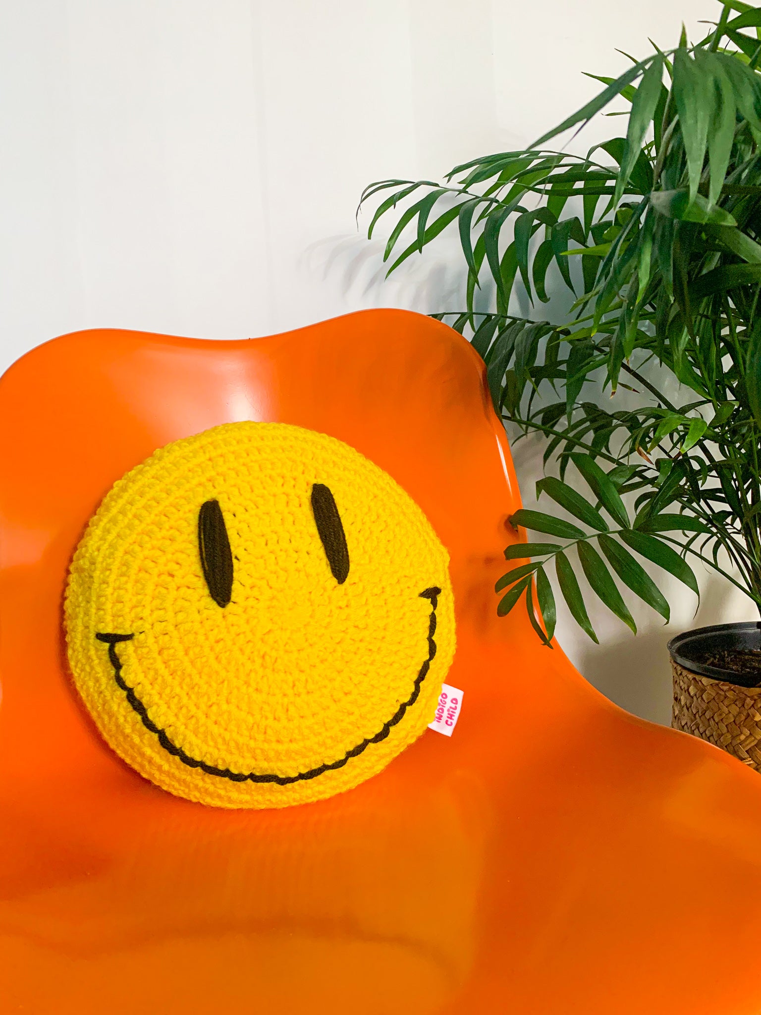 A circle shaped crochet pillow in yellow with a happy face on the front sitting on an orange chair with a plant next to it.