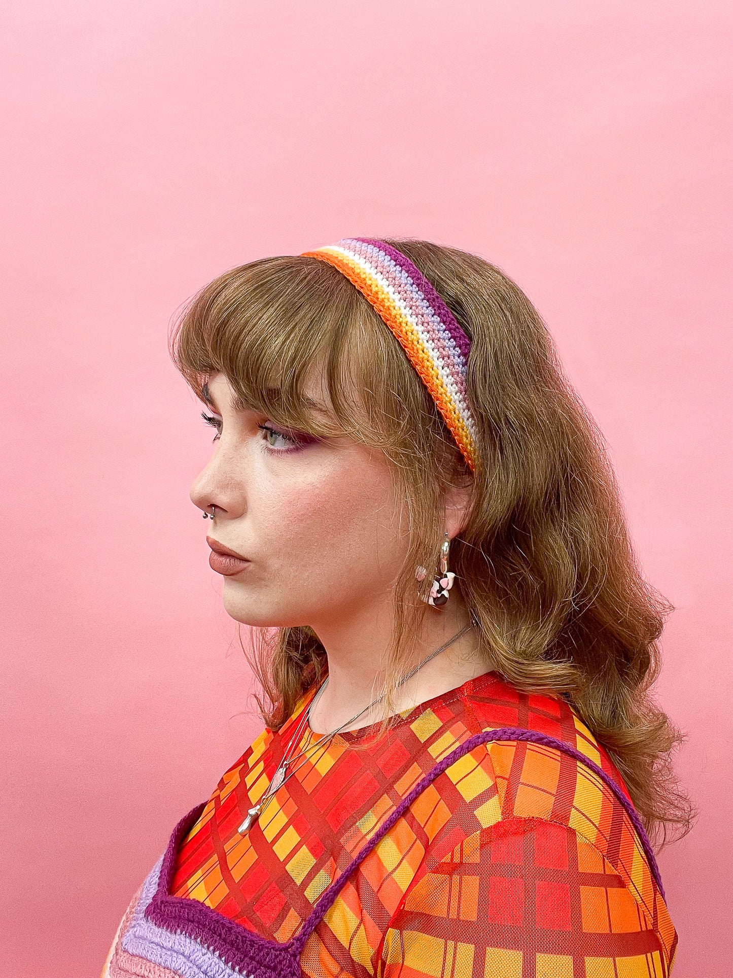 Side view of a woman wearing a crochet headband in the lesbian flag colours (orange, white, pink, purple), with a matching top and orange mesh top underneath.