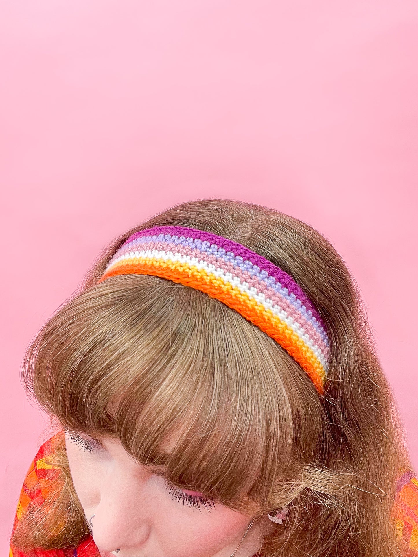 Above angle of a woman wearing a crochet headband with a lesbian flag design in her hair sat in front of a pink background.