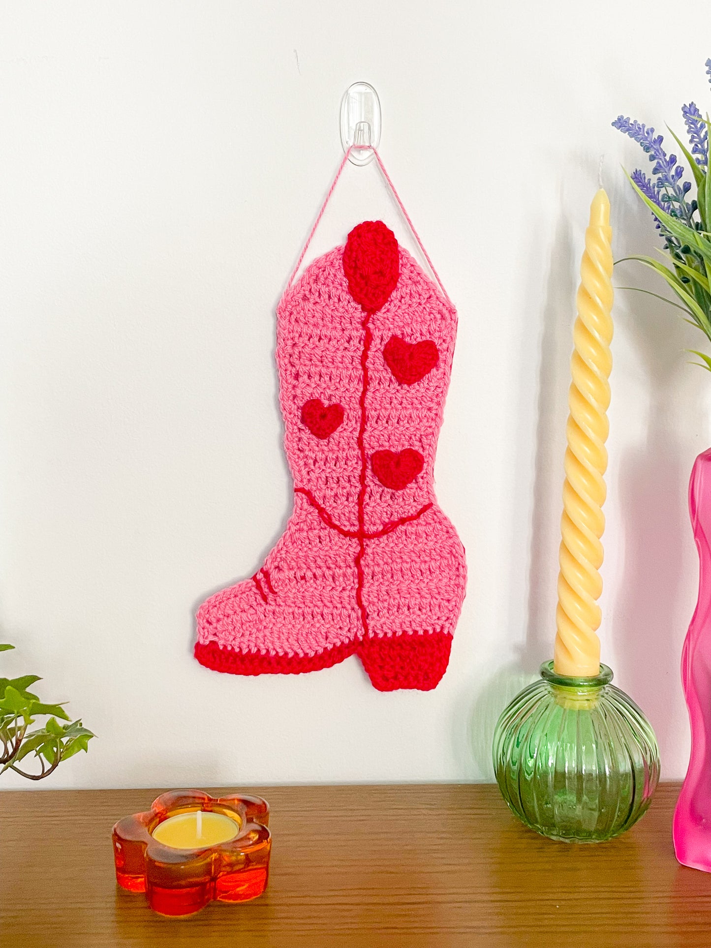 A cowboy boot shaped tapestry in pink with red stitches and detailing hanging on a white wall with candles and plants around it.