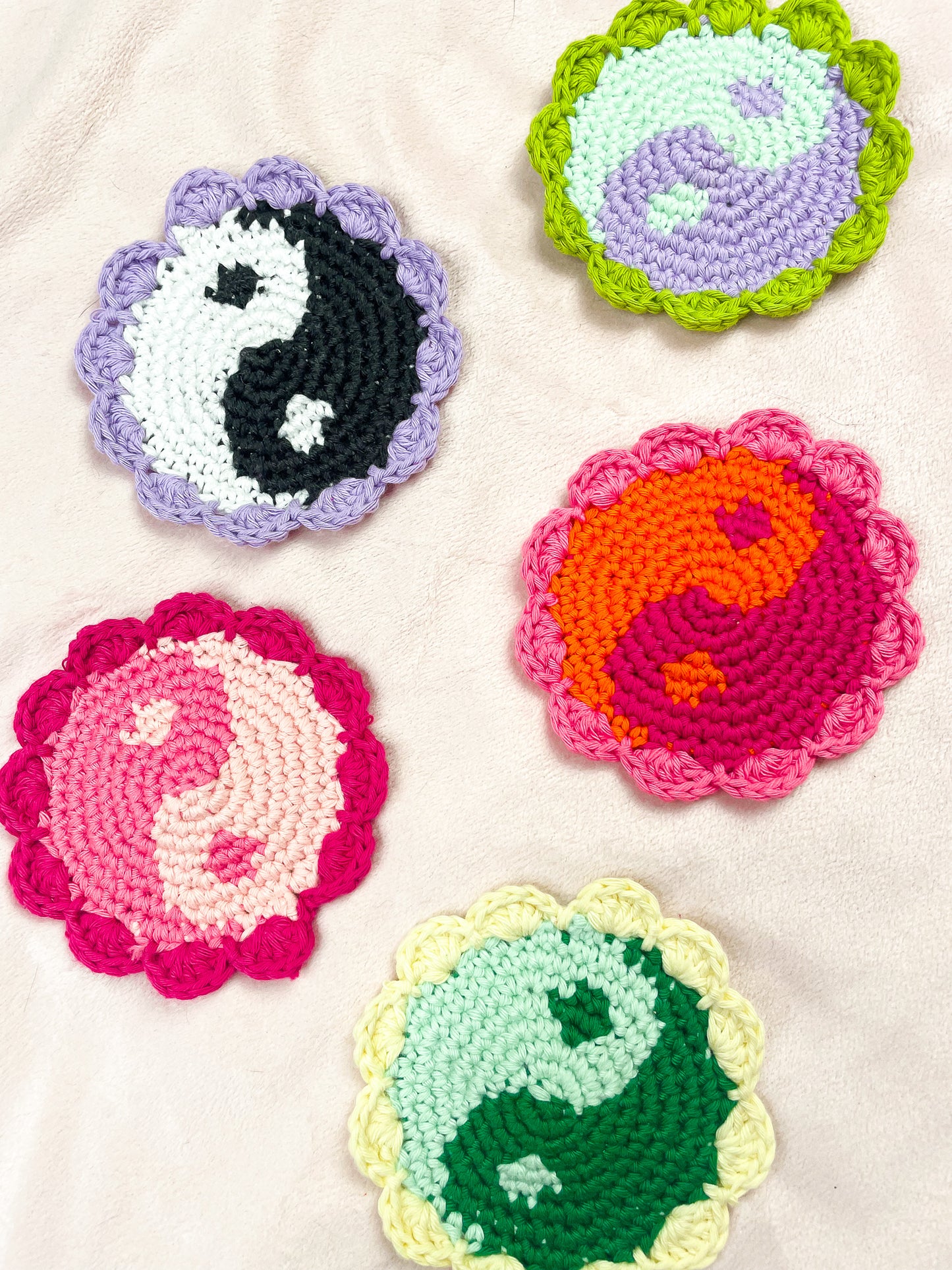 Flat lay of five crocheted coasters with yin yang designs in different colours lying together on a cream background.