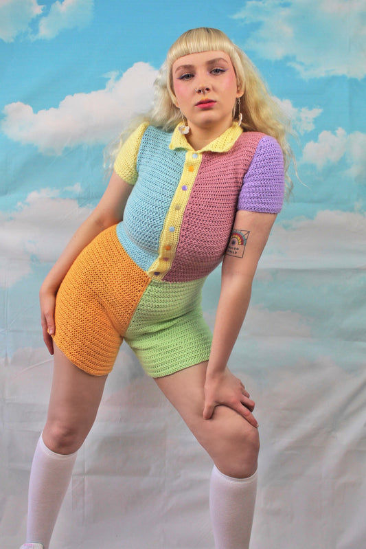 Below view of a woman posing wearing a crochet playsuit in patchwork pastel colours and multicoloured snap buttons standing in front of a cloud background.