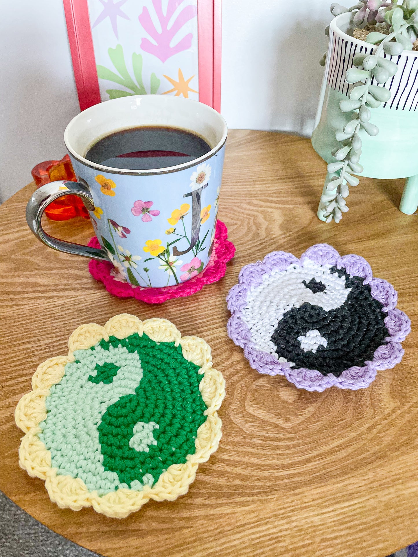 Three crocheted coasters with yin yang designs lying on a wooden coffee table with a coffee cup on one of them and home decor around.