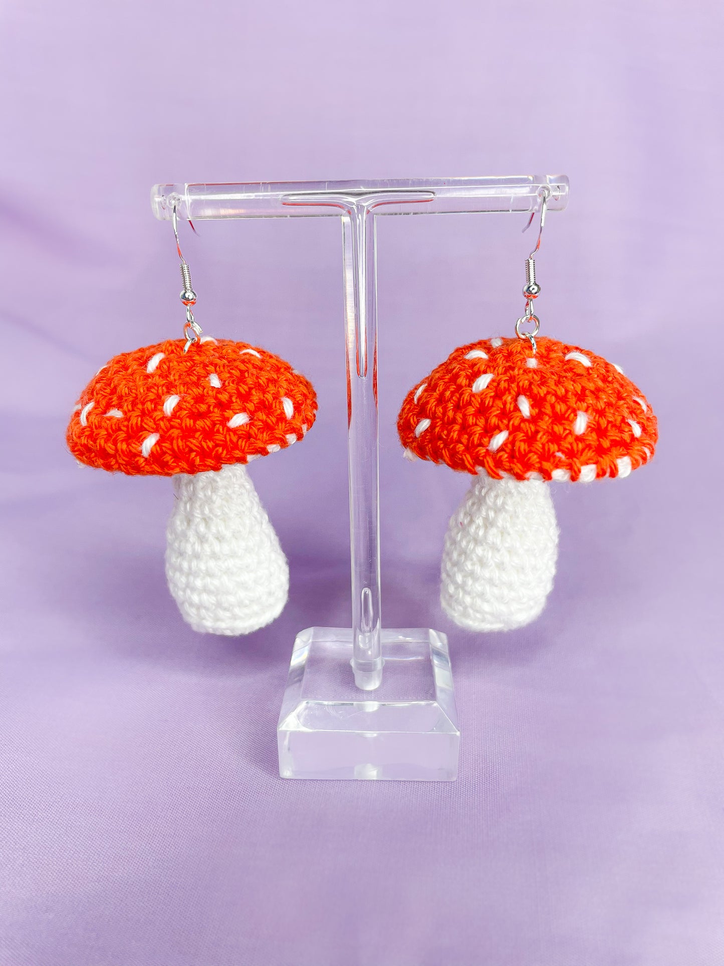 Above view of a pair of orange crochet earrings shaped like mushrooms hanging on an earring stand.