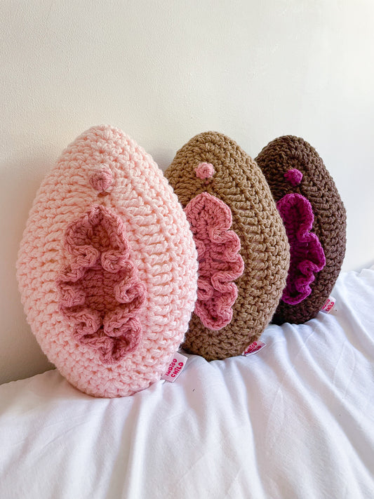 Three crochet pillows leaning together against a wall in a row, one cream, one light brown and the other dark brown, all resembling vulvas.